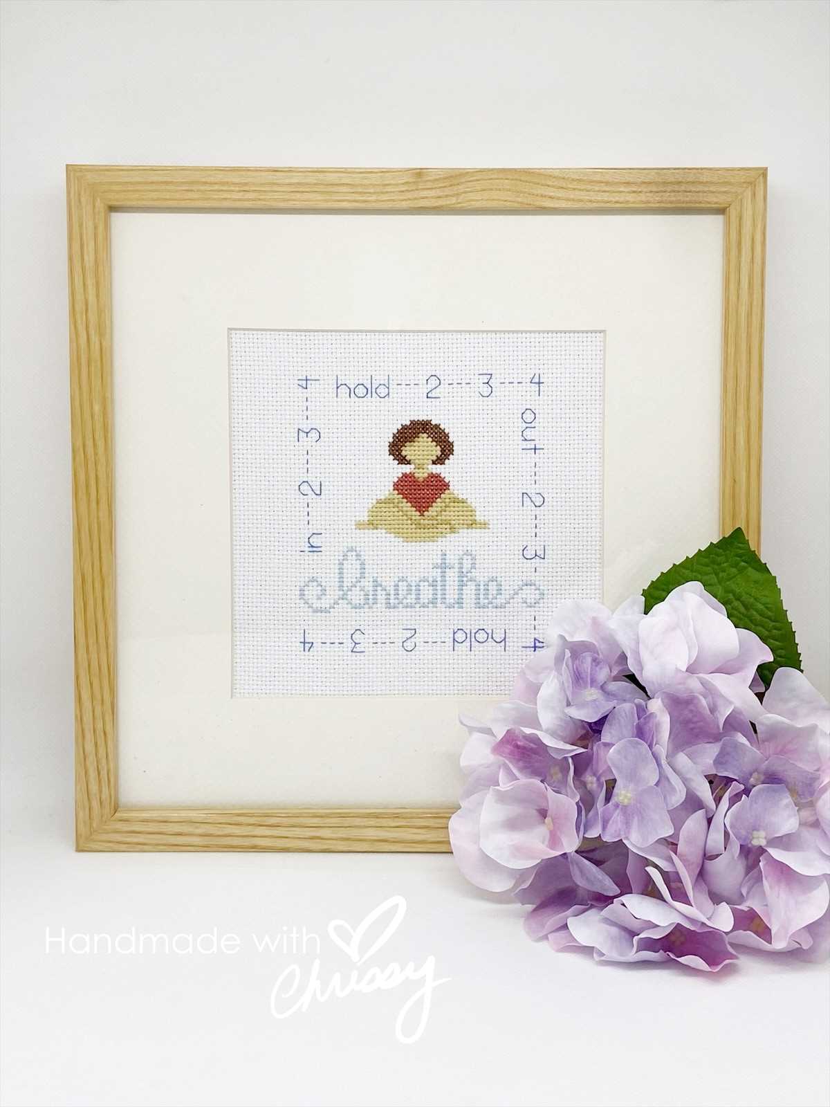 Breathe meditative cross-stitch design by Chrissy Habblett, shown in an oak frame on a white background with a purple flower